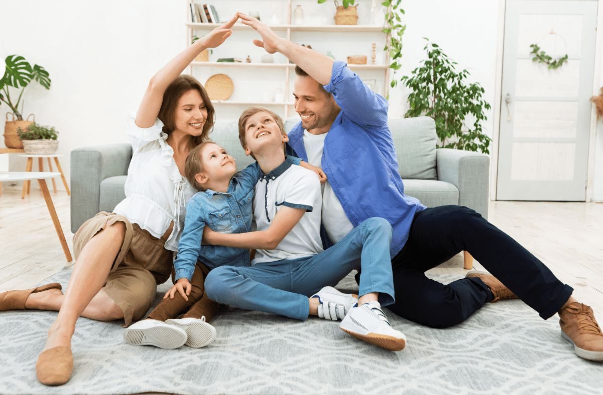 Family sitting on living room floor with mom and dad making a house shape with arms over their two children
