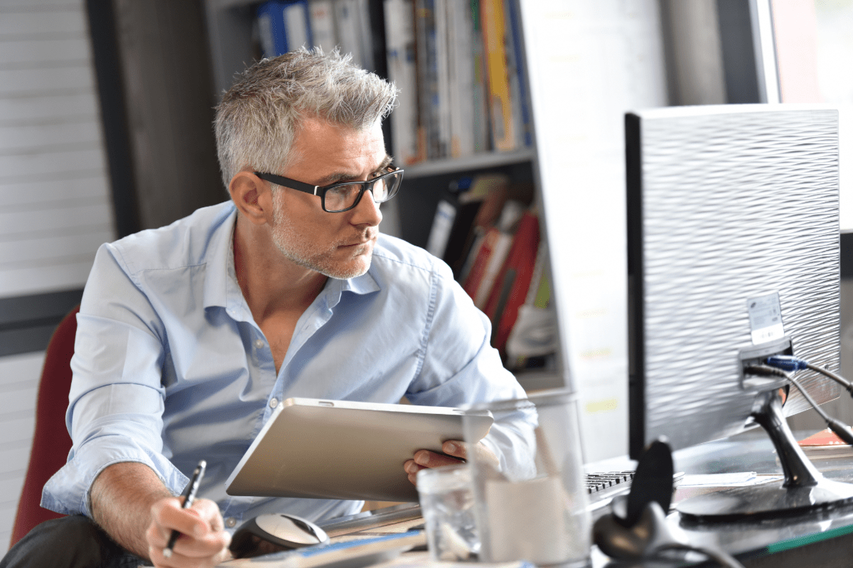 Businessman ditting at desk reviewing insurance documents on tablet and computer