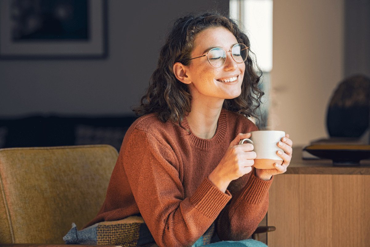 Young adult woman sitting on couch, smiling, drinking from a coffee mug.