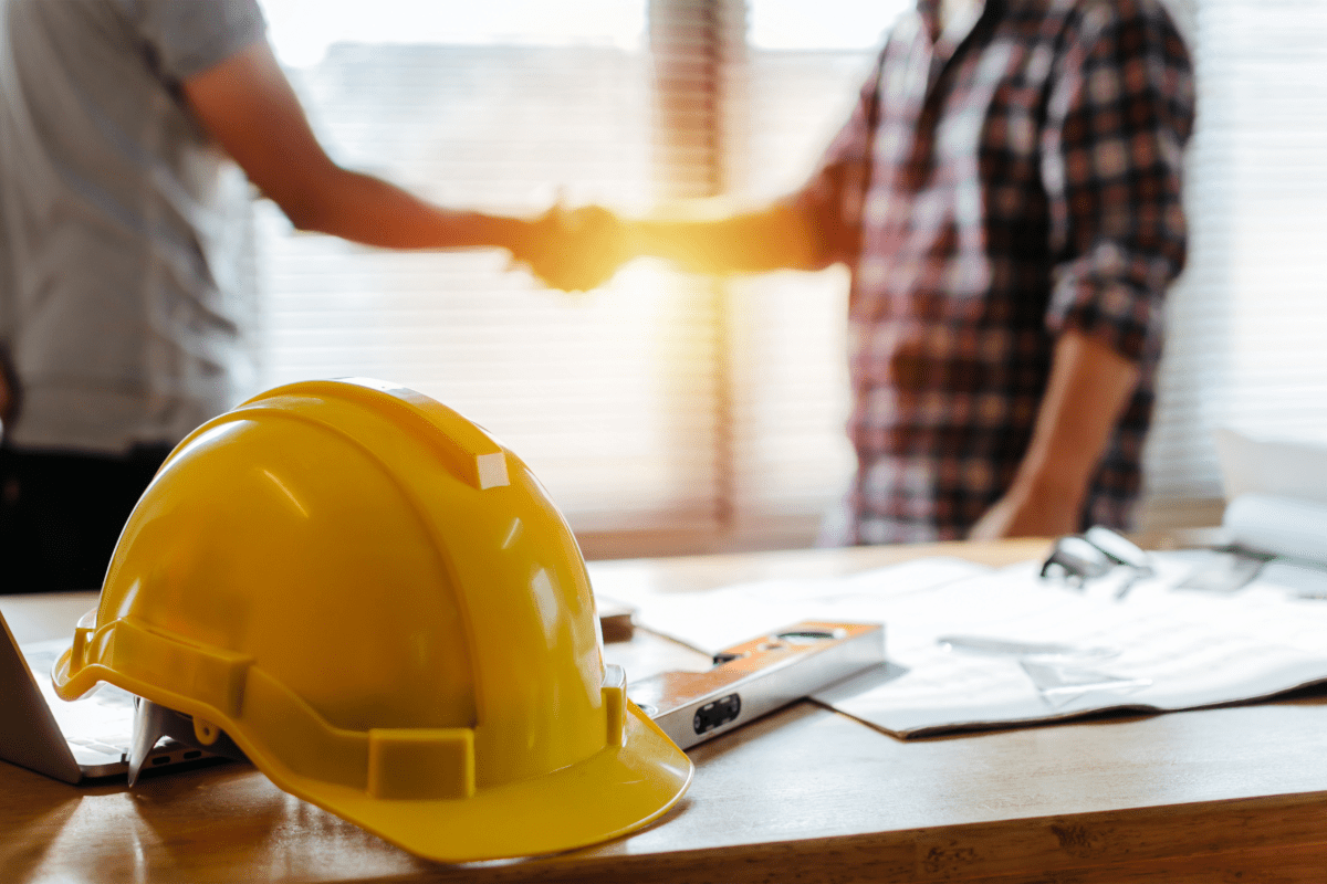 Contractor shaking hands with homeowner with a table with a hard hat and constructions plans in the foreground.