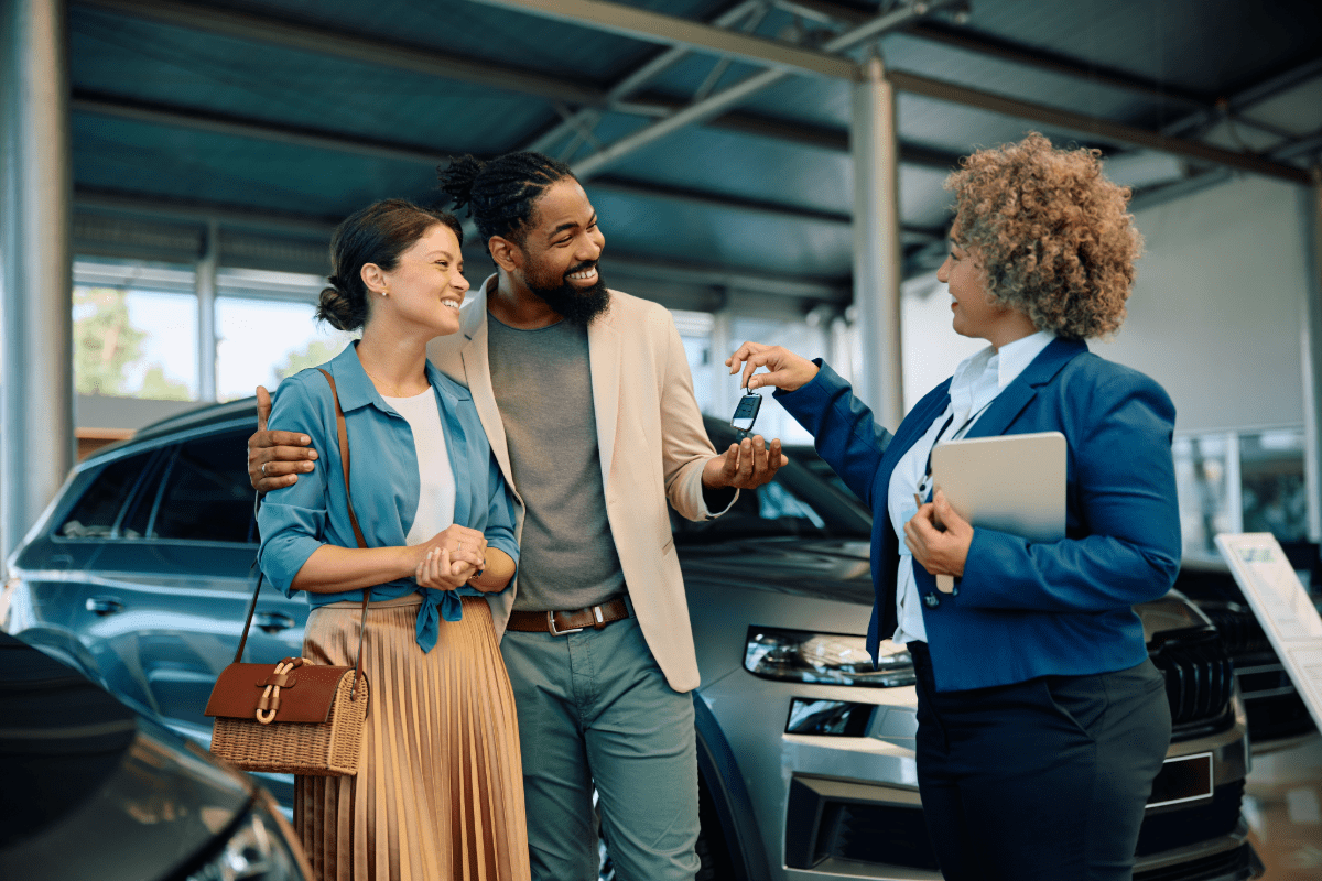 New car saleswoman hands keeps to newly purchased vehicle to a couple.